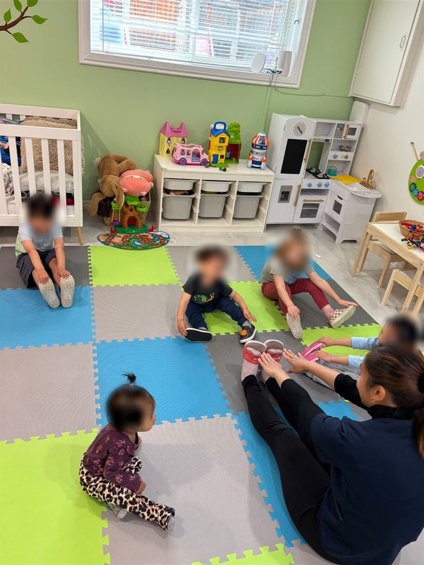 Teacher Marge sitting on a foam play mat leading a group of children with faces blurred for privacy in a seated stretch