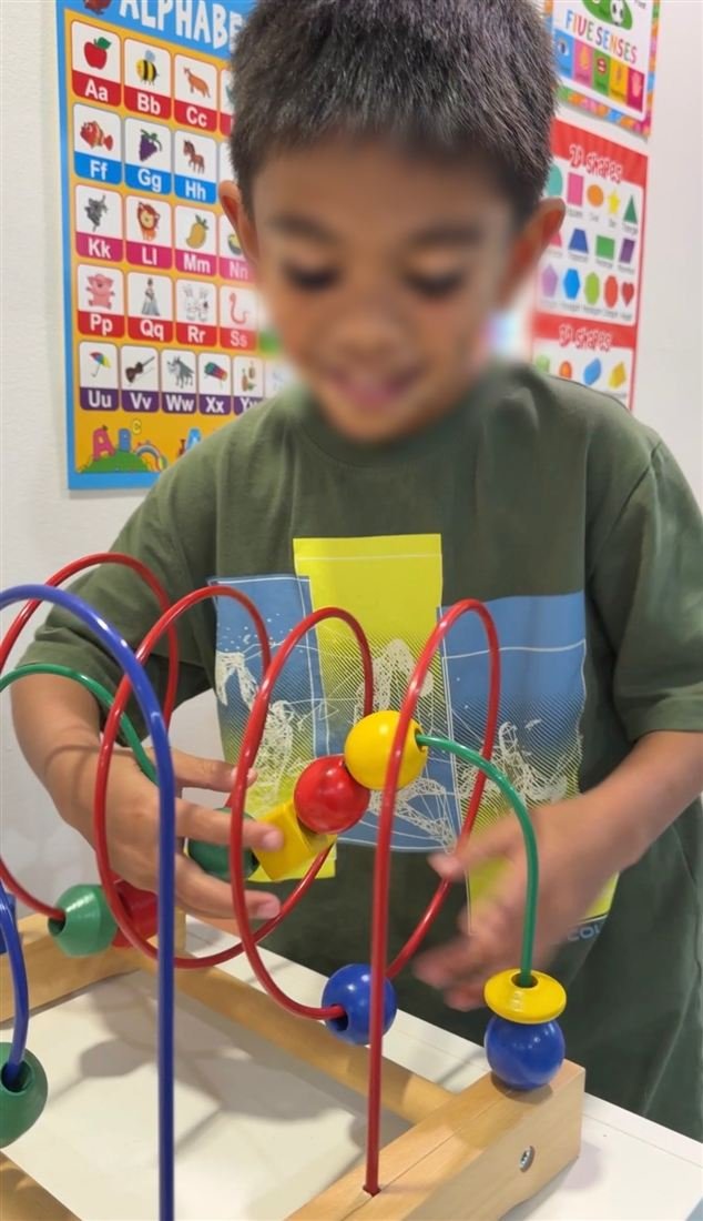A child with a face blurred for privacy sliding colourful beads along a wooden bead maze, with alphabet and shapes posters behind