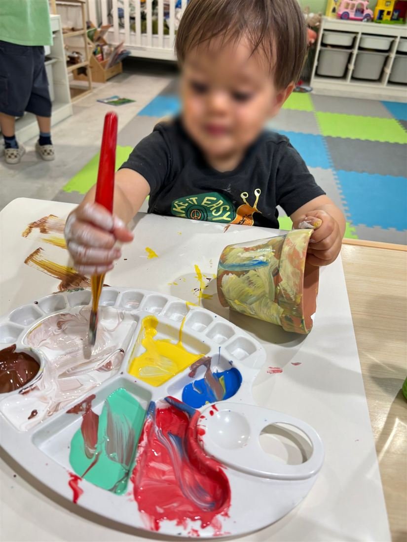 A toddler with a face blurred for privacy painting with a brush and a palette of red, yellow, blue and green paint
