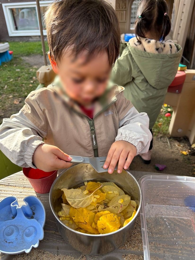A toddler with a face blurred for privacy exploring yellow autumn leaves and corn kernels in a metal pot with a fork