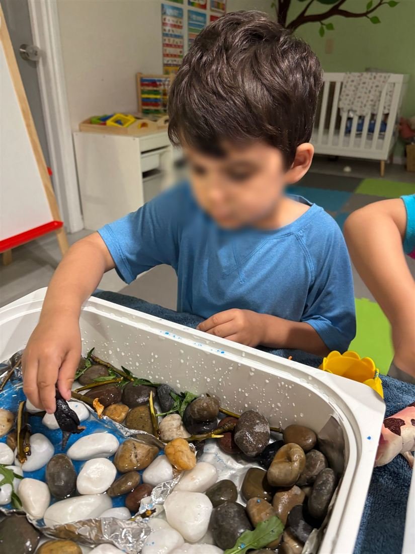 A child with a face blurred for privacy placing a toy penguin among river stones, twigs and blue sand in a white tray
