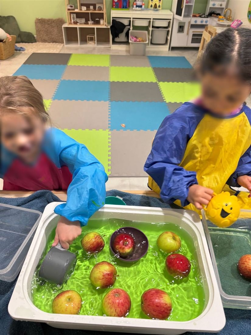 Two children in smocks with faces blurred for privacy playing with apples and cups in a tray of bright green water