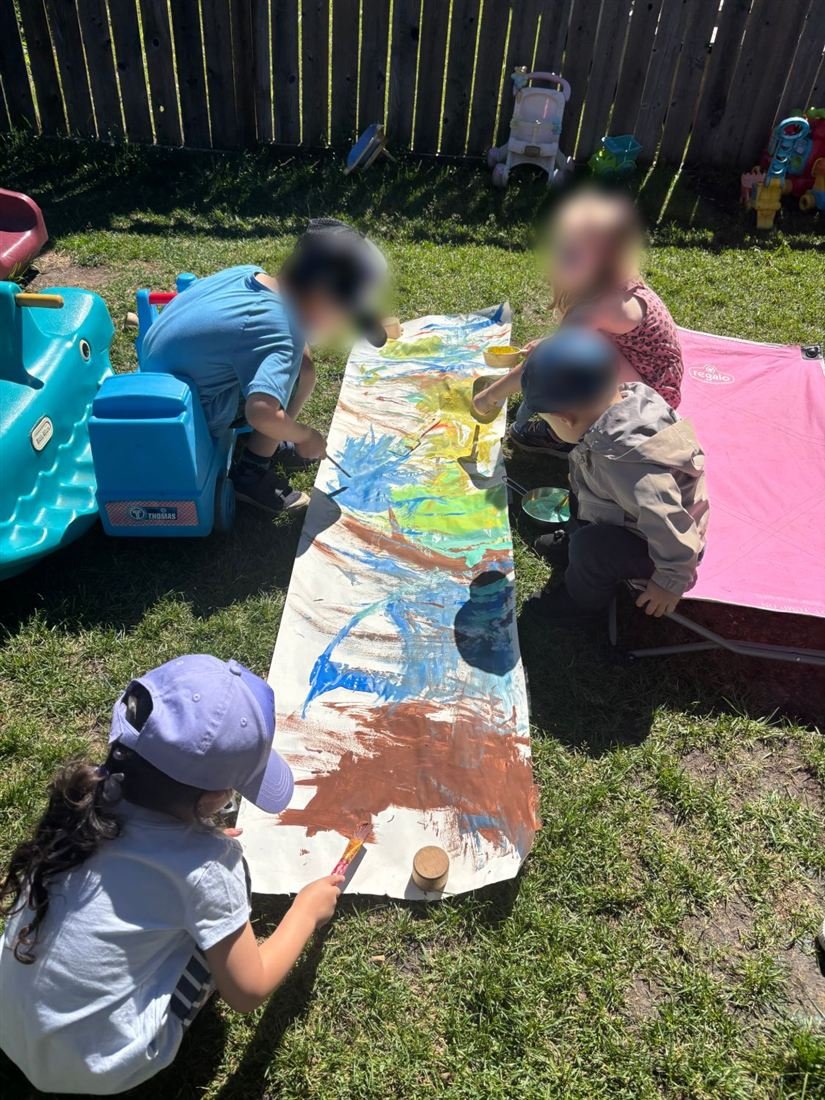 Four children with faces blurred for privacy painting together on a long roll of paper laid out on the backyard grass