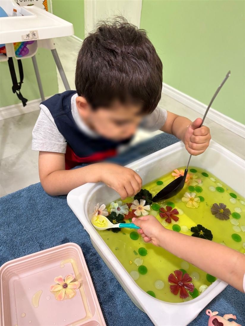 A child with a face blurred for privacy scooping with a ladle in a white bin filled with flowers, green gems and yellow water