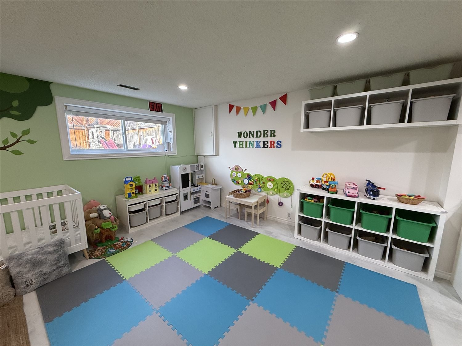 The playroom wall with Wonder Thinkers lettering, colourful bunting, open shelves with bins, a play kitchen and a foam play mat