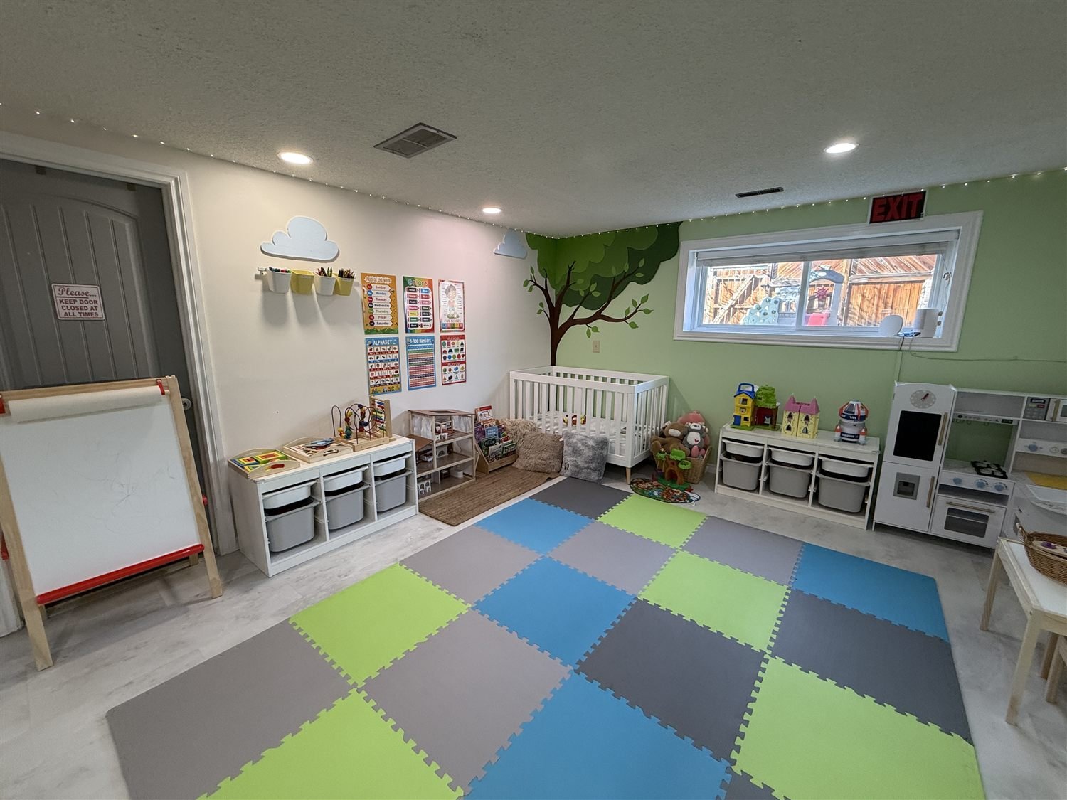 The learning corner with a painted tree mural, learning posters, a crib, a rolling easel and low shelves