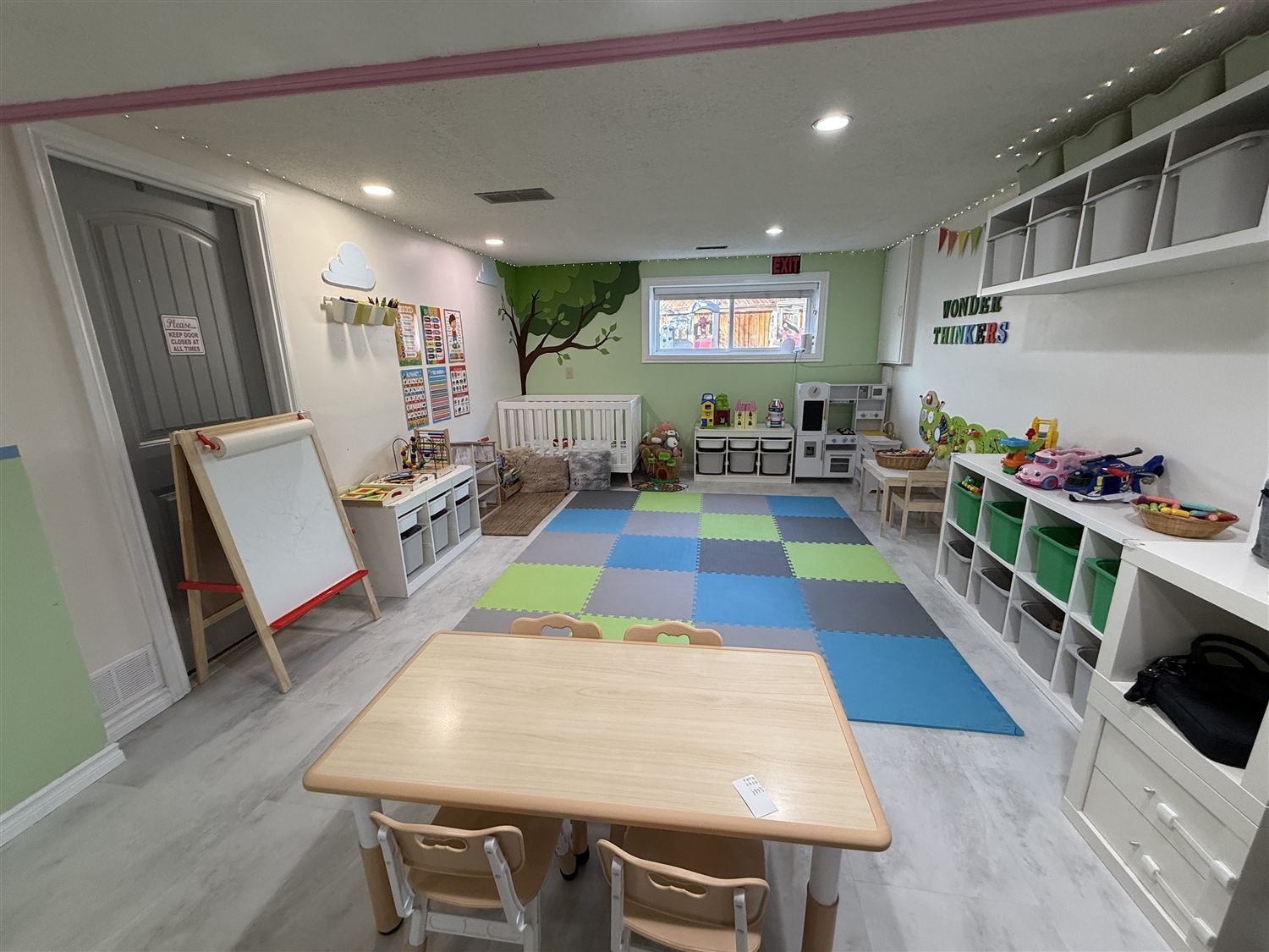 A low wooden table with child-sized chairs in front of an easel, a foam play mat and shelves of toys