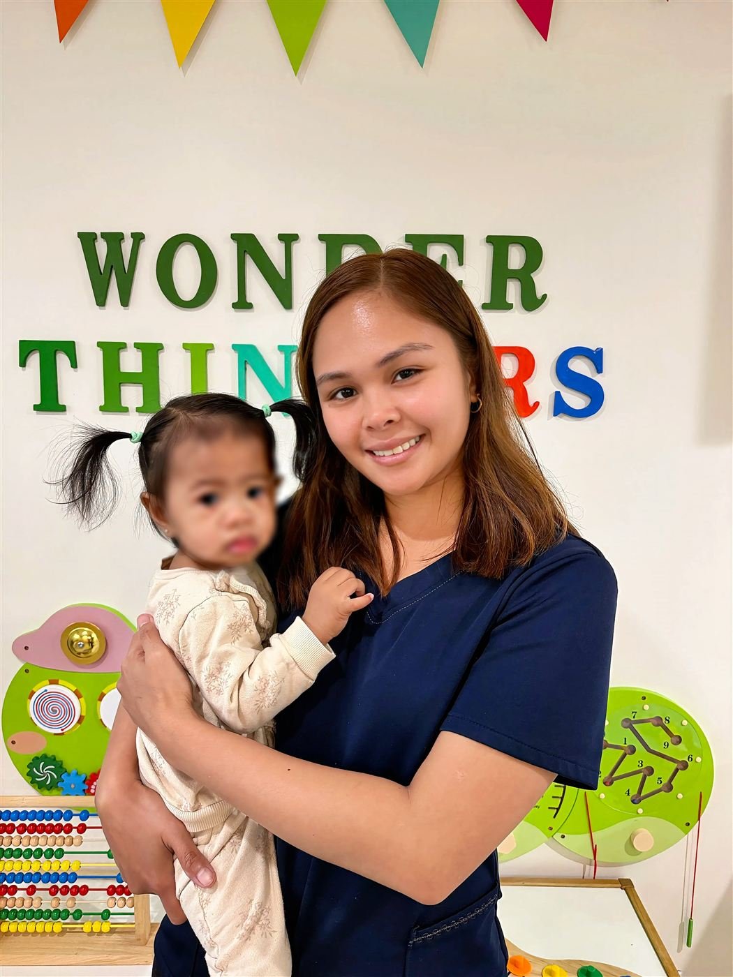 Teacher Marge smiling while holding a toddler in front of the Wonder Thinkers wall lettering