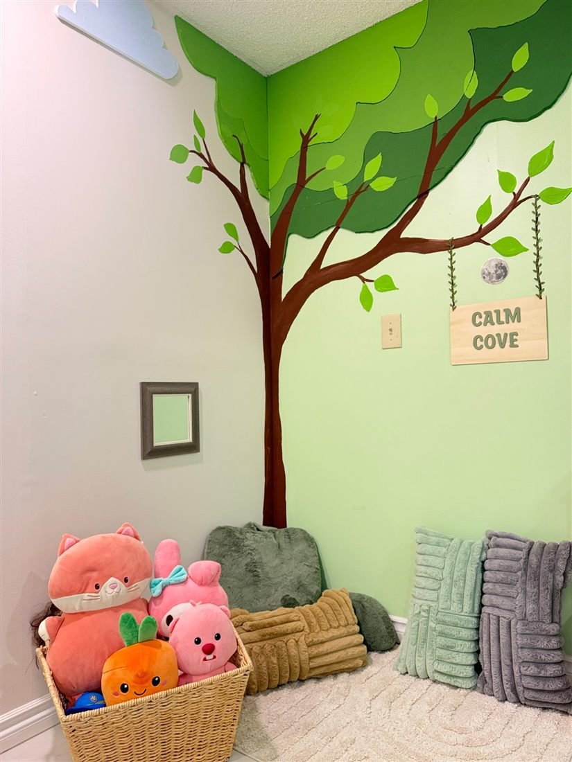 The Calm Cove corner with a painted tree mural, a hanging Calm Cove sign, soft cushions and a basket of plush toys