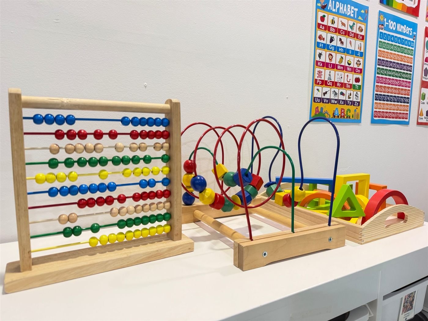 A wooden abacus, a bead maze and a tray of rainbow wooden shape blocks on a low shelf