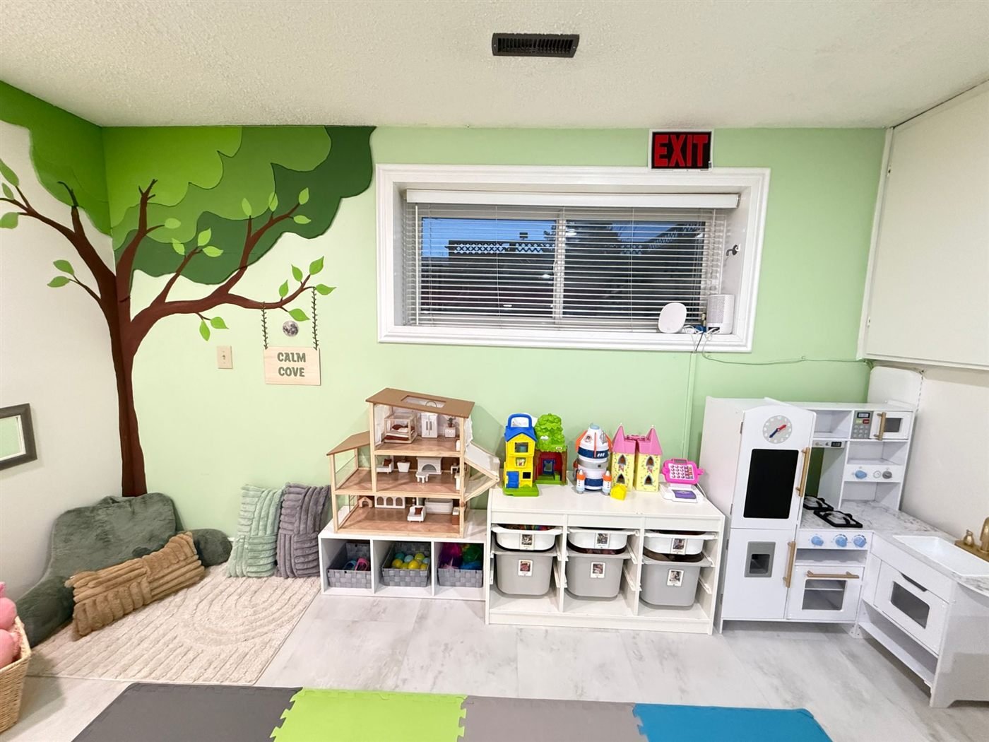 The pretend play area with a wooden dollhouse, toy houses, a play kitchen and a cash register beside the Calm Cove tree mural